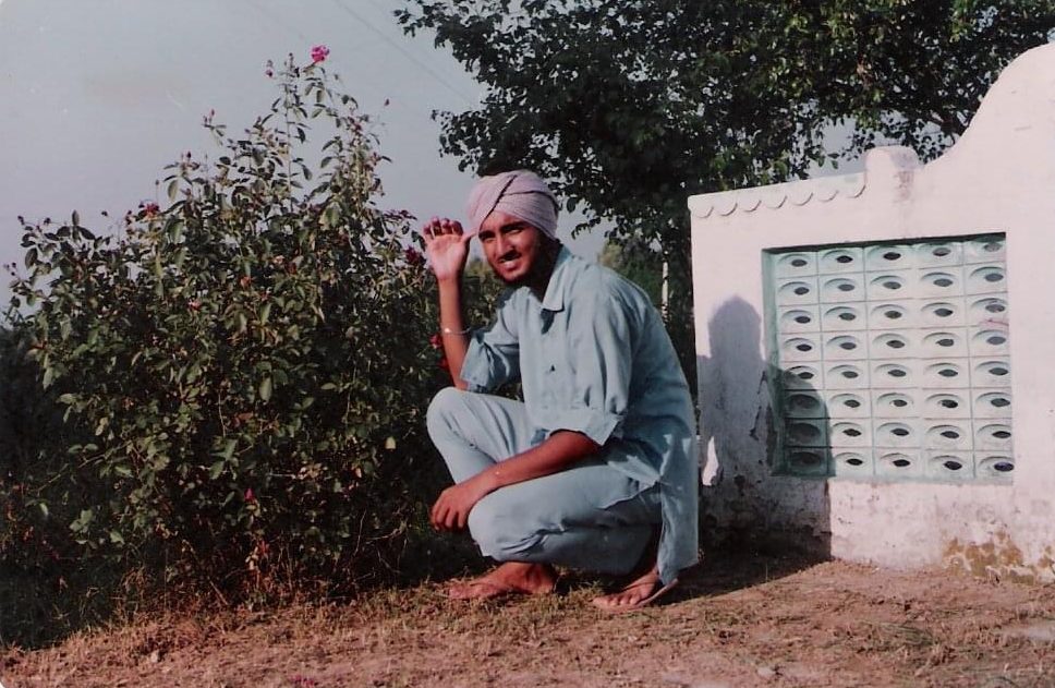 A man wearing a turban is squatting on the ground next to a tree.