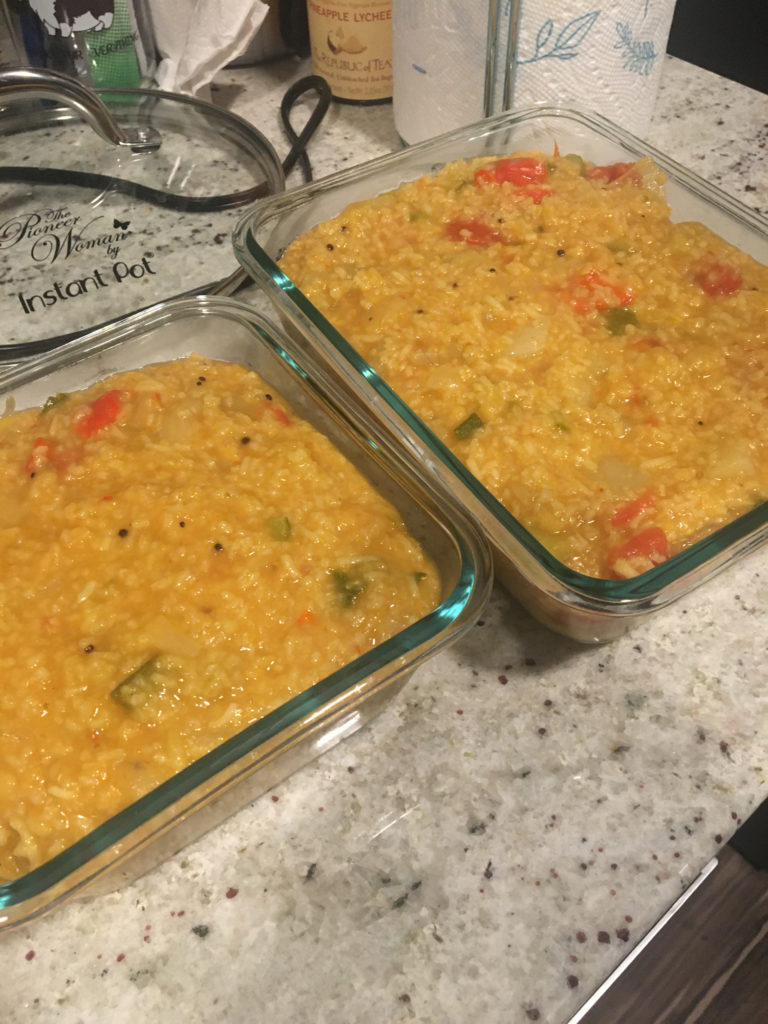 Photograph of finished recipe. Two glass dishes of sambar sadam on a countertop. The thick golden stew is studded with rice, red bell pepper, zucchini, tomatoes and black mustard seeds.